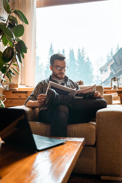 Young adult with glasses and warm clothes reading newspapers while sitting indoors. Man spending his free time alone in the mountains.