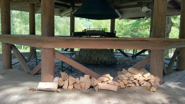 A large wooden gazebo with firewood, a tandoor and a barbecue for outdoor cooking on a sunny summer day. Cooking kebabs on the barbecue for companies of people. Open summer kitchen for preparing barbe