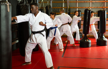 Fototapeta premium African-american man and group of people in kimono exercising with punching bags in gym during karate training.