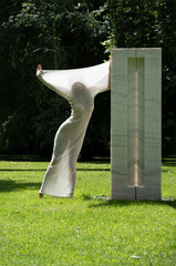 sexy nude woman in white transparent cotton stocking dress as a modern living sculpture outdoors in green nature park backlight at a white marble stone block