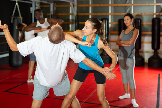 Caucasian man and woman training elbow strike move during self-defence training in gym. Asian woman trainer standing behind and watching.