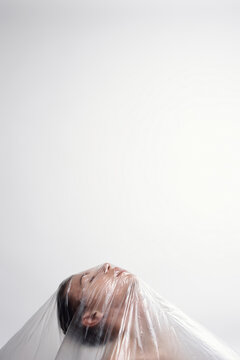 Conceptual headshot of young man standing under polythene sheet with his head up on white wall background    