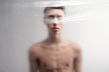Conceptual portrait of shirtless young man with sensual lips standing behind plastic film with his eyes closed 