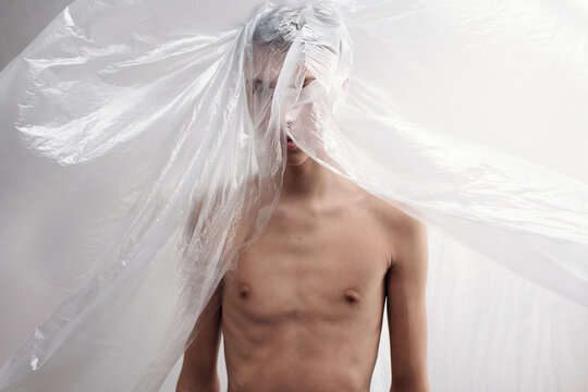 Waist-up portrait of naked young man standing against white wall with face hidden behind polythene sheet   