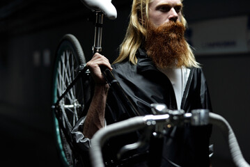 Middle-aged man with long blond hair and full ginger beard walking down the street at night and carrying bike with one hand   