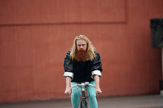 Long-haired blond bearded man in rain jacket riding a bicycle along the street on rainy day against brown wall background 