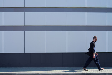 Side view of young man in casual clothes walking by modern office building with glass facade  