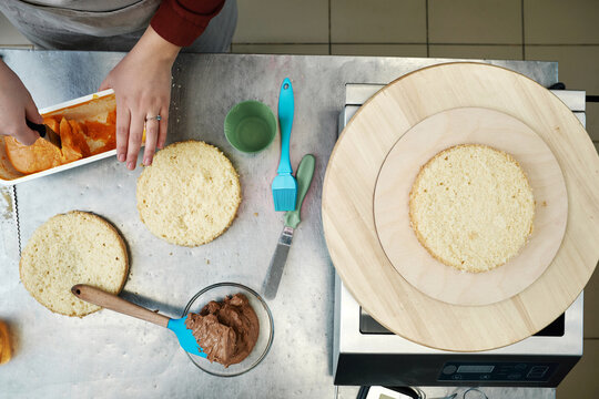 Directly above view of unrecognizable female cook taking mango filling from container and getting ready to spread it on cake layer   