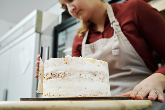 Low angle view of young professional female confectioner coating sponge cake with whipped cream frosting in kitchen 