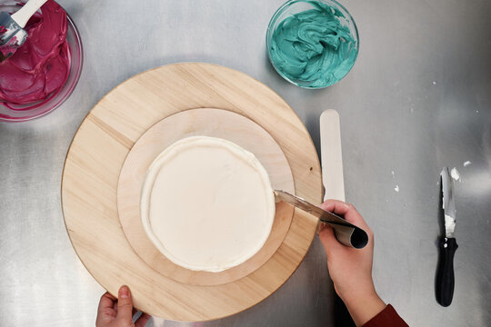 Top view of unrecognizable pastry chef coating cake with white cream, bowls with colorful toppings seen on table beside 