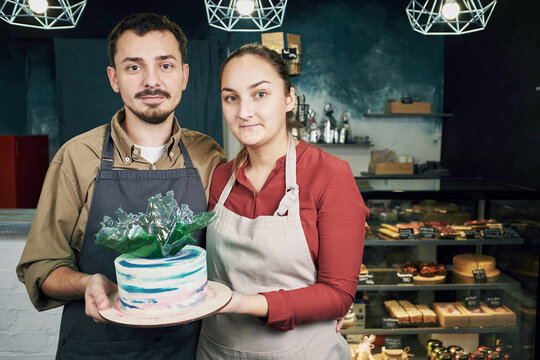Naklejki Portrait of young male and female pastry cooks standing in confectionery, holding beautiful cake with sugar glass decoration and looking at camera 