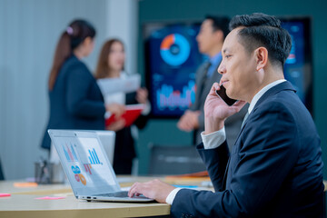 Smiling middle-aged executive asian businessman sitting at a worktable a modern office, typing on computer keyboard, and talk to work, sending emails to his business partners, working on marketing.