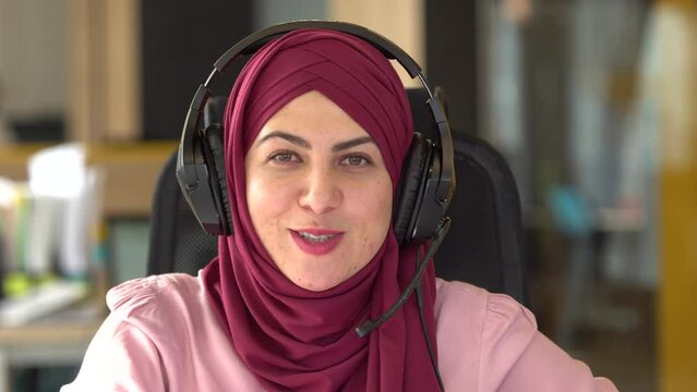 Portrait of cheerful muslim girl wearing hijab looking into the camera and chatting with microphone and headphones online video communication