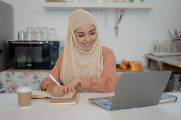 A beautiful Asian Muslim female student is studying for an online class at a table in the kitchen