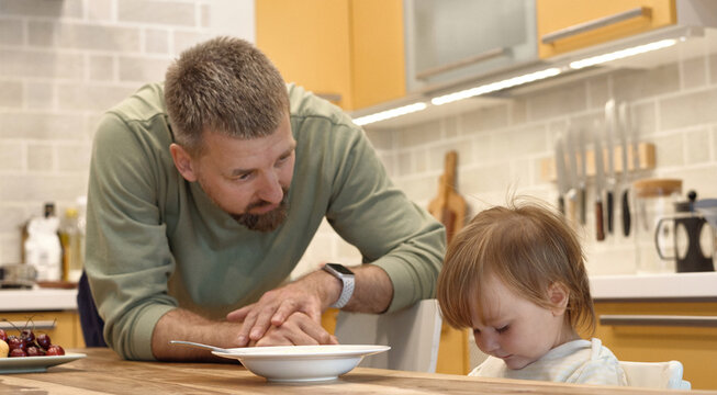 Bearded man trying to feed little child at table in kitchen