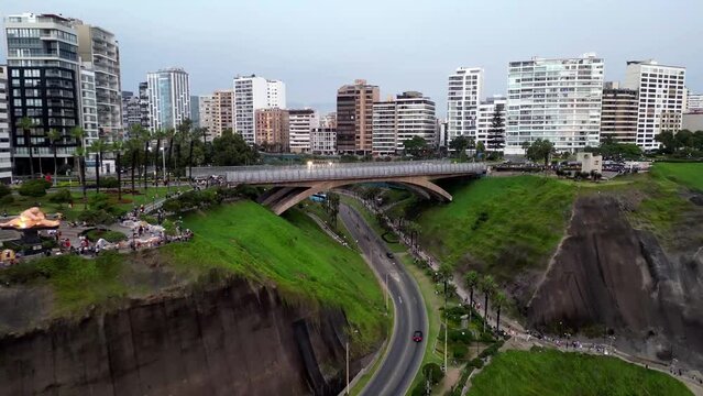 Aerial dolly back shot showing a southamerican concrete city bridge connecting two coast grass ravines surrounding by buildings, parks with palm trees and people walking