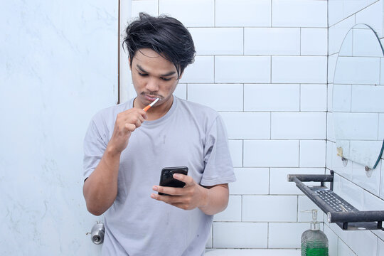 Young Asian man with toothbrush cleaning teeth and looking to mobile phone at the bathroom