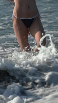 Front view slim unrecognizable woman with long legs in bikini bottom walking on beach knee deep in splashing water of breaking waves Pacific Ocean. Cropped waist down of female. Vertical, slow motion