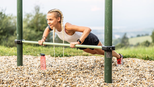 Sporty young female working out calisthenics in an outdoor gym using parallel bars. Beautiful blonde girl with braids works out outdoors doing push-ups on an elevated bar