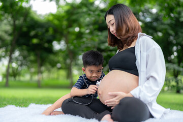 Portrait of a pregnant woman relaxing in the park with her son. Older brother uses headphones to listen to his younger brother in the womb