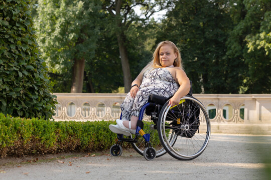 Full Length Young Pretty Woman With Short Stature On Wheelchair Looking At Camera in Public Park At Summer Day. Smiling Pretty Female Adult With Disability. Horizontal Plane