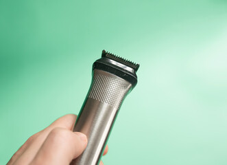 Close up hand of unknown caucasian man holding electric beard trimmer cordless hair cutter on green background.