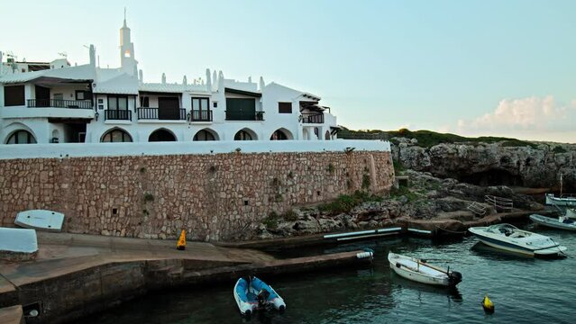 Boats by the seashore of Binibeca Vell with a town view in the background in 4K. Calming sea and early morning scenery in Menorca, Balearic Islands, Spain with whitewashed houses and rocky caves.