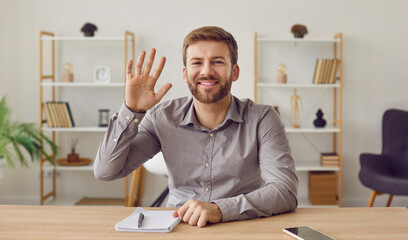 Businessman having video call looking at webcam waving his hand in greeting. Portrait of smiling business leader, speaker sitting at desk looking at camera during webinar, online class, webcam view - obrazy, fototapety, plakaty