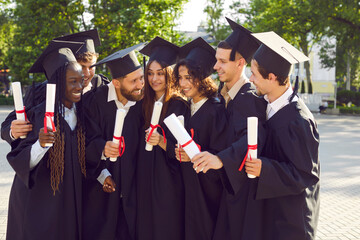 Group of seven college or university students having fun on graduation day. Happy diverse friends in graduate hats and gowns, holding diploma scrolls, standing in green campus yard, talking and joking