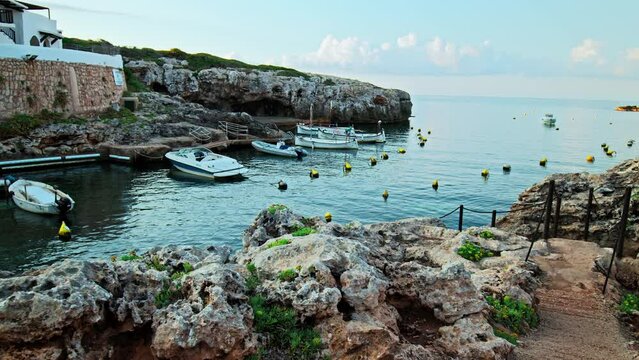 Boats by the seashore of Binibeca Vell with a town view in the background in 4K. Calming sea and early morning scenery in Menorca, Balearic Islands, Spain with whitewashed houses and rocky caves.
