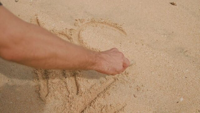 Close-up of a man's hand holding a stick drawing symbols in the sand. A person draws a plan of action on the sand in a schematic form. Summer entertainment on the beach. Drawing on white tropical sand