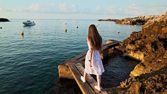 Golden sunrise falling over a beautiful girl crossing a wooden bridge in the sea. A young woman in a white dress walking by the seashore rocky edge enjoying corals in the Mediterranean Sea in Spain.