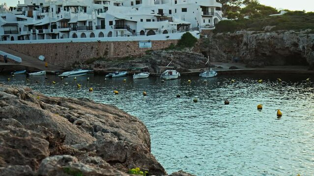 Boats by the seashore of Binibeca Vell with a town view in the background in 4K. Calming sea and early morning scenery in Menorca, Balearic Islands, Spain with whitewashed houses and rocky caves.