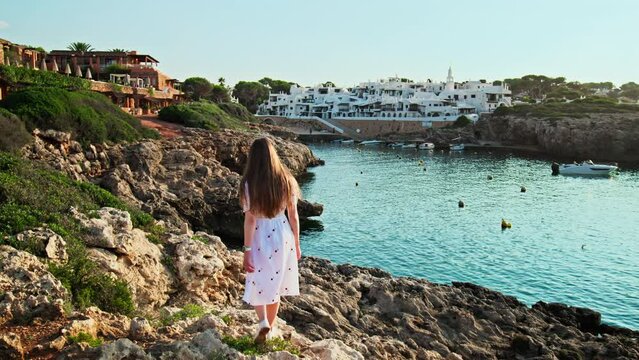 Girl in white dress long hair sitting on a rocky seashore with a beautiful view. Attractive female enjoying nature early in the morning during sunrise in Binibeca Vell, Menorca, Spain