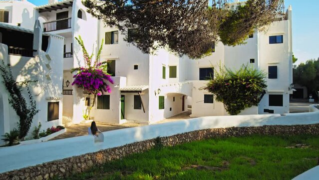Tourist girl enjoying beautiful purple flowers attached to a whitewashed house. A female sitting and watching a colourful bush of flowers growing on a wall in a town resort in Binibeca, Menorca, Spain