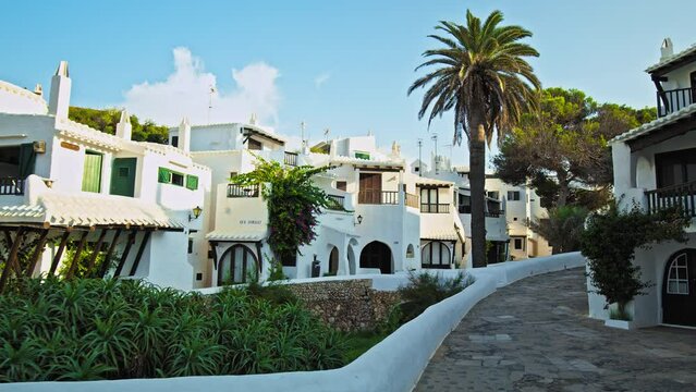 Scenic view of Binibeca Village with whitewashed houses and tropical green trees. Relaxing tourist resort Binibeca Vell with narrow streets and a calm cove - paradise-like town in Menorca, Spain.