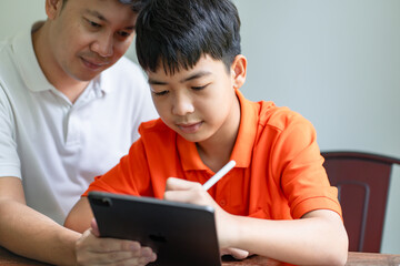 Fototapeta premium Asian father and son using tablet&nbsp;teaching son to draw.playing with digital tablet at desk fatherhood