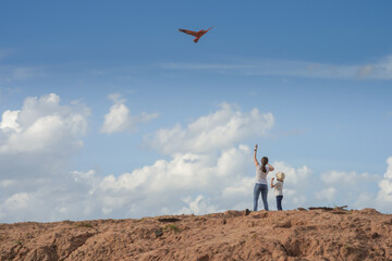 Obraz premium Mother and daughter in jeans and white t-shirts Flying a kite in an earthen field with a blue sky and white clouds in the background.