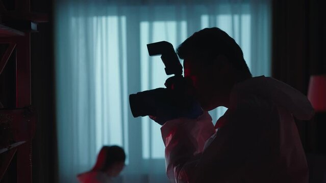 Forensic scientists collect evidence at the crime scene, in a dark apartment lit by blue red light from police sirens. A woman inspects the murder weapon. A man takes pictures with a photo camera.