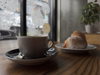A hot cup of coffee is served next to a freshly baked bread roll at a window counter in a cafe.
