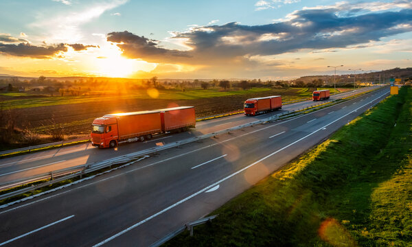 Three orange Transportation trucks with trailers on an asphalt highway road in a rural landscape at sunset
