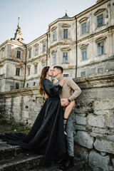 Fototapeta na wymiar Woman in black long dress hugs man stand on stairs near ancient palace. Female kissing male in street. Luxury couple newlyweds near old Pidhirtsi Castle, Lviv, Ukraine. Stylish bride and groom outdoor