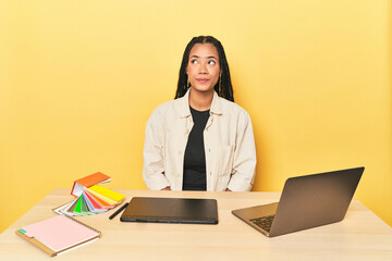 Indonesian designer with tools at table on yellow excited keeping ok gesture on eye.