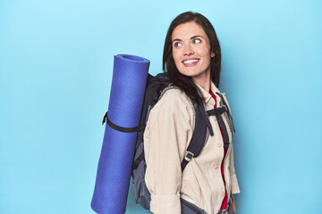 Adventurous woman with backpack on blue looks aside smiling, cheerful and pleasant.