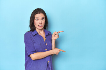 Young caucasian woman on blue backdrop shocked pointing with index fingers to a copy space.