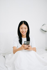 Beautiful young asian woman on a white bed with coffee cub and using smart phone at home. lifestyle concept