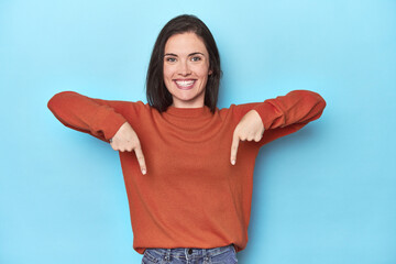 Young caucasian woman on blue backdrop points down with fingers, positive feeling.