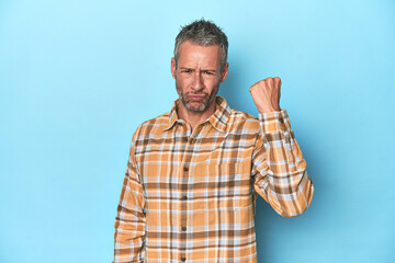 Fototapeta premium Middle-aged caucasian man on blue backdrop showing fist to camera, aggressive facial expression.