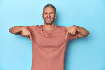 Middle-aged caucasian man on blue backdrop points down with fingers, positive feeling.