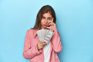 Teen girl in pink, flaunting a handful of dollar bills pointing temple with finger, thinking, focused on a task.